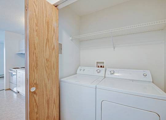 A convenient in-home laundry area here at Aspen Terrace with a full-size washer and dryer and a wide overhead wire shelf for storage. A nearby doorway opens to the kitchen, creating an easy, functional layout.