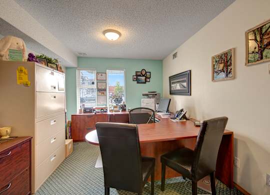 A welcoming office area here at Aspen Terrace with a spacious wood desk, guest chairs, and natural light from front windows. A tidy workspace with filing cabinets, computer setup, and warm wall décor creates a professional, friendly feel.