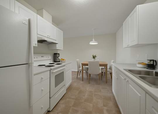 A spacious kitchen and dining area here at Aspen Terrace with white cabinets, matching white appliances, and a double stainless steel sink on wide countertops. A simple dining table with upholstered chairs sits under a modern pendant light, giving the room a clean, bright feel.