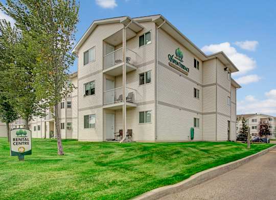 A sunny exterior view here at Aspen Terrace featuring a well-kept apartment building with private balconies and freshly landscaped green lawns. A rental center sign and nearby parking create a welcoming, convenient community setting.