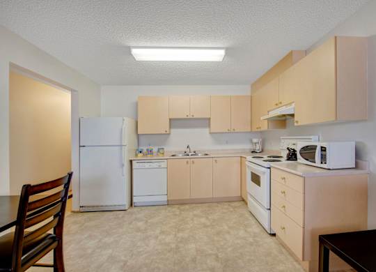 A spacious kitchen here at Aspen Terrace with light wood cabinets, white countertops, and matching white appliances including a refrigerator, stove, and dishwasher. A double sink and open floor space create a clean, bright layout with a simple, neutral feel.