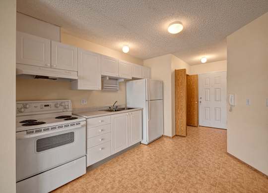 A bright kitchen here at Aspen Terrace with white cabinets, white appliances, and a stainless steel sink with generous counter space. A wide tiled floor and overhead lighting create an open, practical layout with a clean, neutral feel.