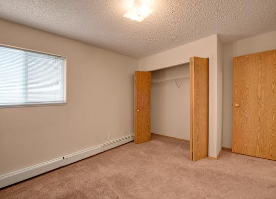 A bright, empty bedroom here at Aspen Terrace with beige carpeting, a wide window with blinds for natural light, and a spacious closet with wood sliding doors. A clean, open layout creates a comfortable, move-in-ready feel.