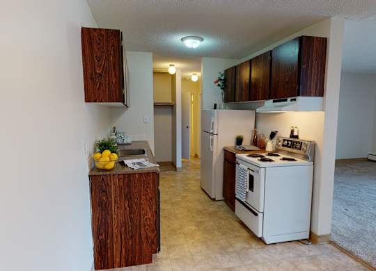 A clean kitchen area here at Cambridge West with dark wood cabinets, a white refrigerator, and a stove with bright overhead lighting. A long countertop with a sink, small decor accents, and a bowl of lemons adds a welcoming touch, with the space opening into the carpeted living room nearby.