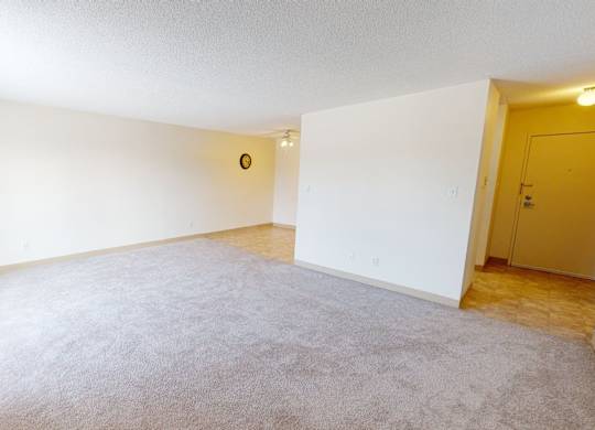 A bright open living room here at Cambridge West with soft carpet flooring, crisp white walls, and a wide layout with plenty of open space. Large windows with vertical blinds bring in natural light, while the entry hallway with tile flooring and warm overhead lighting adds a practical flow into the home.