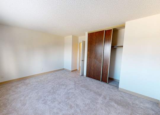 A bright, empty bedroom here at Cambridge West with soft beige carpet and light neutral walls, featuring a large window for natural light and a spacious closet with sliding wood doors for convenient storage.