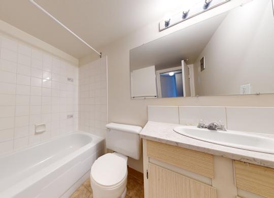 A clean bathroom here at Cambridge West with a bathtub-shower combo, white tiled walls, and a white toilet, featuring a light vanity with a spacious countertop and a large mirror under bright lighting.