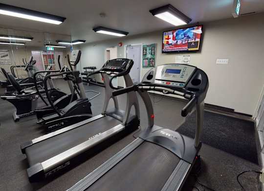 A well-equipped fitness room here at Cambridge West with treadmills, cardio machines, and free weights, featuring mirrored walls, rubber flooring, and bright lighting with a mounted TV for added convenience.