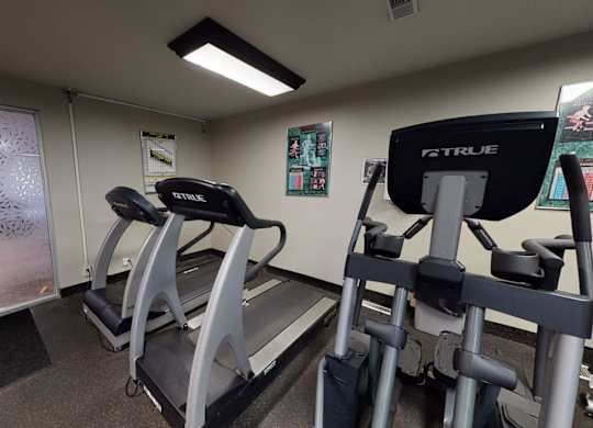 A modern fitness room here at Cambridge West with treadmills and cardio equipment, featuring rubber flooring, bright overhead lighting, and easy access to the community lounge through glass doors.
