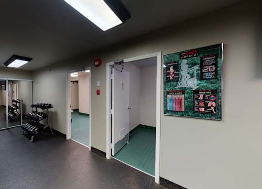 A clean fitness center hallway here at Cambridge West with rubber flooring, bright overhead lighting, and a free weight rack, offering easy access to workout areas and nearby community spaces.