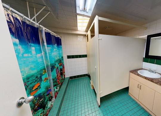 A clean shared bathroom here at Cambridge West with tiled floors, colorful shower curtains, and private shower stalls, featuring a double-sink vanity with a spacious countertop and a large mirror under bright lighting.