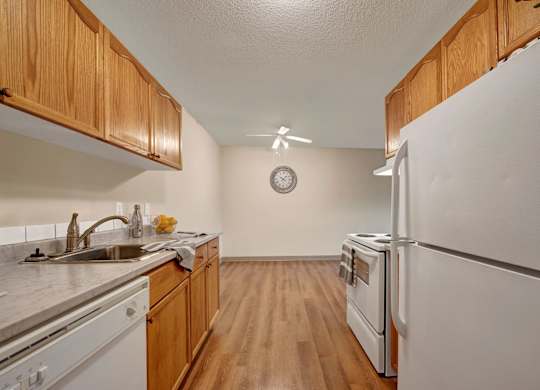A bright kitchen and dining space here at Cambridge West with warm wood cabinets, white appliances, and a long countertop with a stainless sink and dishwasher. Wood-style flooring, a ceiling fan, and an open dining area with a wall clock create a simple, functional layout with plenty of room to move around.