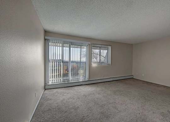 A bright living room here at Cedar Gardens with a wide open layout and soft carpeting, featuring large sliding glass doors with vertical blinds, an extra window for added natural light, and a long baseboard heater along the wall for a cozy, comfortable feel.