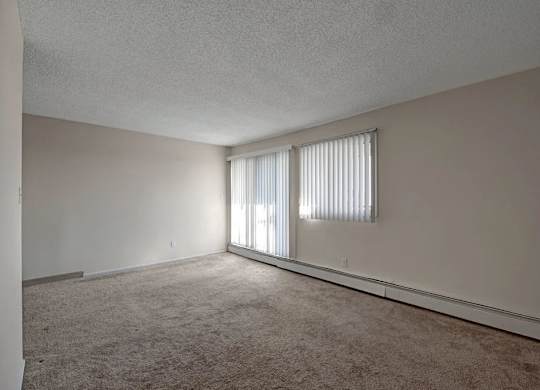 An airy living room here at Cedar Gardens with plush carpeting and a long open layout, featuring a sliding glass door with vertical blinds, an additional window for natural light, and a baseboard heater along the wall for a warm, comfortable feel.