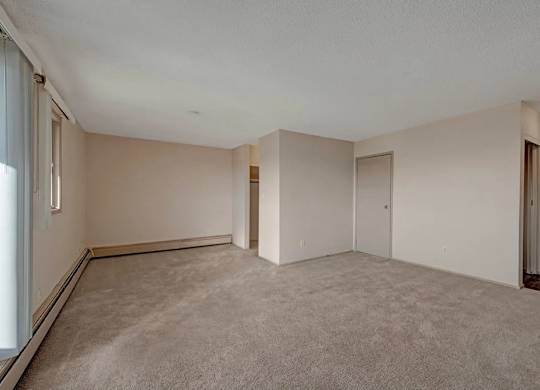 A spacious living area here at Cedar Gardens with soft carpeting and a bright, open layout, featuring a large window with vertical blinds, a long baseboard heater along the wall, and a versatile nook near the entryway for a cozy, comfortable feel.