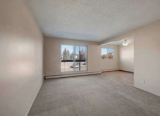 A bright open living room here at Cedar Gardens with soft carpeting and a wide layout, featuring a large sliding glass door for natural light, a baseboard heater along the wall, and a connected dining nook with an additional window and ceiling fan for an airy, comfortable feel.