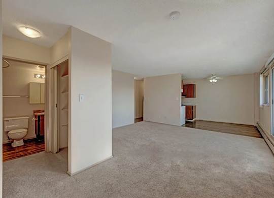 A spacious open living area here at Cedar Gardens with soft carpeting and an easy flow into the kitchen space, featuring large sliding glass doors with wide windows for natural light, a long baseboard heater, and nearby built-in shelving and bathroom access for added everyday convenience.