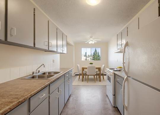 A bright kitchen here at Cedar Gardens with plenty of cabinet storage and generous counter space, featuring a double sink, white appliances, and an open dining area with a cozy table set by the window for a clean, comfortable feel.