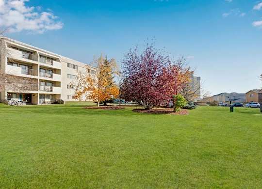 A spacious outdoor courtyard here at Cedar Gardens with wide open green space and mature trees, featuring colorful landscaping, a peaceful lawn area for relaxing or playing, and clear views of the surrounding community buildings under a bright blue sky.
