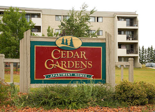 Here at Cedar Gardens, a welcoming entrance sign with red and green accents sits in landscaped greenery and rock mulch, with a multi-story apartment building, balconies, and mature trees in the background.