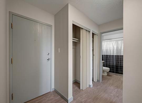 A bright entryway here at Cedar Gardens with wood-style flooring and a sleek front door, featuring convenient closet storage with sliding doors, clean neutral walls, and a simple hallway layout that leads easily into the bathroom for an open, welcoming feel.