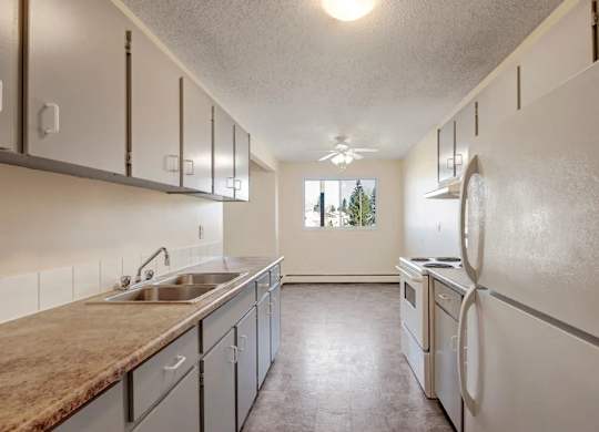 A clean kitchen here at Cedar Gardens with sleek white cabinetry and long countertops, featuring a double-basin sink, full-size appliances, and bright ceiling lighting that leads into the dining space for a simple, comfortable feel.