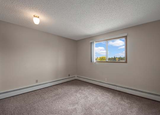 A bright, empty bedroom here at Gateway Square with soft beige walls, plush carpet flooring, and a large window with vertical blinds letting in natural light and peaceful outdoor views.