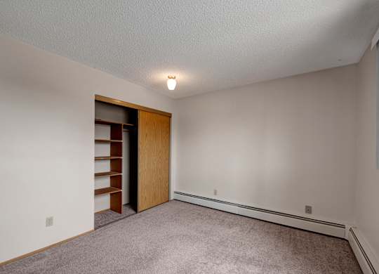 A spacious, empty bedroom here at Gateway Square with soft beige walls, plush carpet flooring, and a large closet featuring sliding wood doors and built-in shelving for convenient storage.