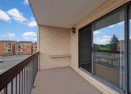 A private outdoor balcony here at Gateway Square with durable flooring, a sliding glass door leading inside, and open views of nearby buildings and greenery under a bright sky.