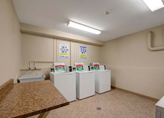 A clean, well-lit laundry room here at Gateway Square with multiple top-loading washers, neutral walls, durable flooring, and a spacious folding counter for added convenience.