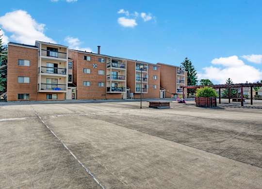 A well-maintained apartment building here at Gateway Square with private balconies, warm-toned exterior siding, open courtyard space, and landscaped planters under a bright blue sky.