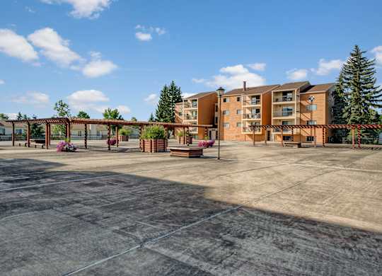 A spacious outdoor courtyard here at Gateway Square with open concrete walkways, wooden pergolas, landscaped planters, and surrounding apartment buildings under a bright blue sky.