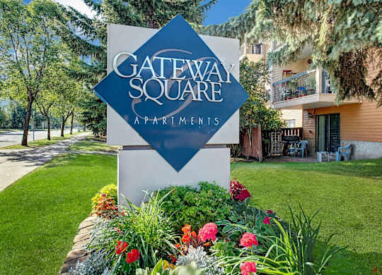 A welcoming community entrance sign here at Gateway Square surrounded by manicured landscaping, colorful flowers, mature trees, and nearby apartment buildings in a calm residential setting.