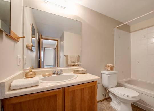 A bright bathroom here at Gateway Square featuring a wide vanity with a large wall mirror and a clean countertop sink, paired with warm wood cabinetry, soft neutral walls, and a classic tub-shower combo for a fresh, comfortable space.