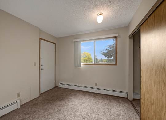 A bright, empty bedroom here at Gateway Square featuring soft neutral walls, plush carpet flooring, and a large window with vertical blinds that fills the space with natural light, plus a spacious closet and simple layout for a comfortable retreat.