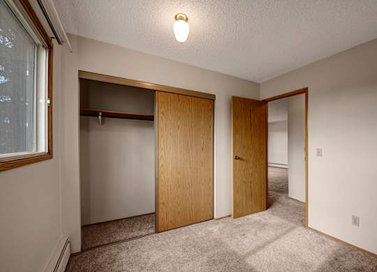 A bright, empty bedroom here at Gateway Square featuring soft neutral walls, plush carpet flooring, and a large window with warm wood trim, plus a spacious sliding-door closet and an open layout that feels clean and comfortable.