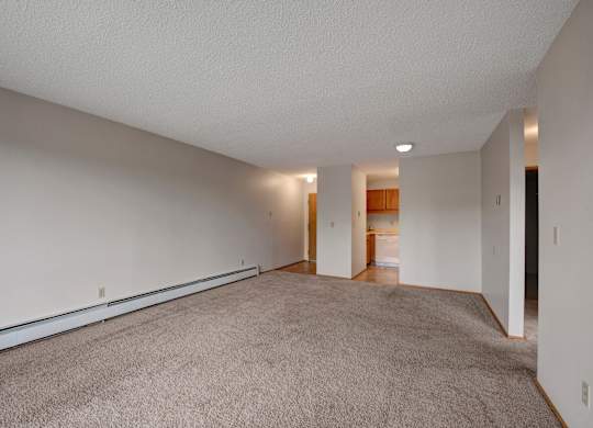 A bright open living space here at Gateway Square featuring soft neutral walls, plush carpet flooring, and a long layout that offers plenty of room to relax, with a cozy flow leading into the kitchen area for easy everyday living.