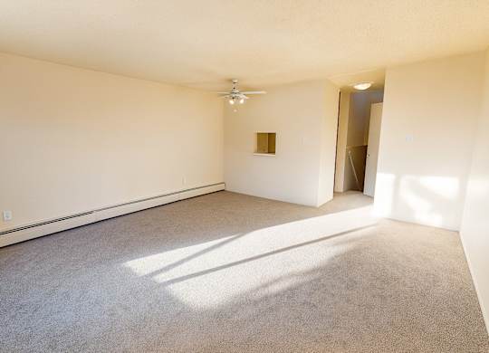 A sunlit living room at Lakewood Heights featuring soft carpet, neutral walls, baseboard heating, ceiling fan lighting, and an open, flexible layout with a pass-through detail that adds natural flow and everyday comfort.