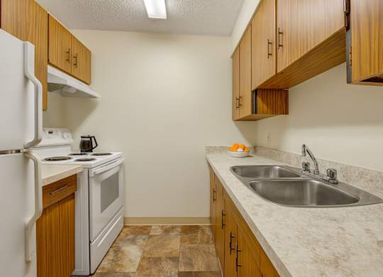 A clean, functional kitchen here at Lakewood Heights with warm wood cabinets, light countertops, a stainless steel double sink, white appliances, and efficient galley-style layout with bright overhead lighting.