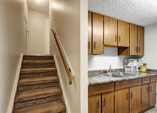 A practical interior at Lakewood Heights showcasing a carpeted staircase with handrail alongside a functional kitchen featuring wood cabinetry, ample storage, durable countertops, and bright overhead lighting.