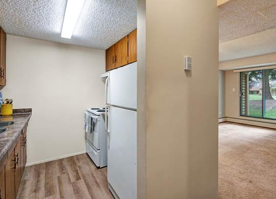 A bright kitchen and living area at Lakewood Heights featuring wood cabinetry, durable flooring, efficient appliances, and an open layout that flows into a carpeted living space with a large sliding glass door for natural light.