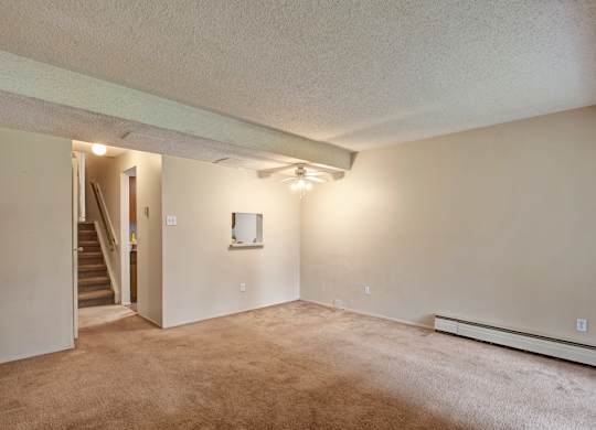 A spacious living room at Lakewood Heights with plush carpet, neutral walls, exposed ceiling beam detail, ceiling fan lighting, and an open layout that flows easily toward the stairway and adjacent spaces.