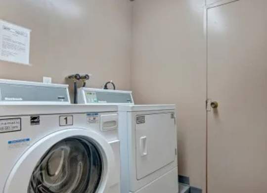 Laundry room here at Madison Park Apartment Homes with a white front-loading washing machine and dryer against a beige wall. Notice sign on the wall adds a utilitarian feel.