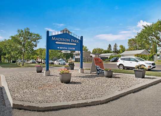A welcoming community entrance sign here at Madison Park with a large blue monument sign displaying the apartment name and rental center direction. Decorative planters with flowers sit on a gravel landscape bed, while nearby streets, parked vehicles, mature trees, and neighboring homes appear under a bright, clear sky.