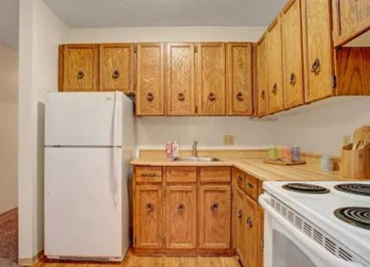 Small kitchen here at Madison Park Apartment Homes with a white fridge, wooden cabinets, and a beige countertop. Includes a sink, stove, and neatly arranged kitchen items, conveying a cozy feel.
