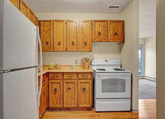 A cozy kitchen here at Madison Park Apartment Homes features wooden cabinets, a white refrigerator, and a sink. Countertops hold utensils, cups, and plates, creating a warm, inviting atmosphere.