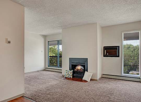 A cozy living room here at Madison Park Apartment Homes with a carpeted floor and a central fireplace framed by pillows. Large windows on each side let in natural light and offer views of greenery.