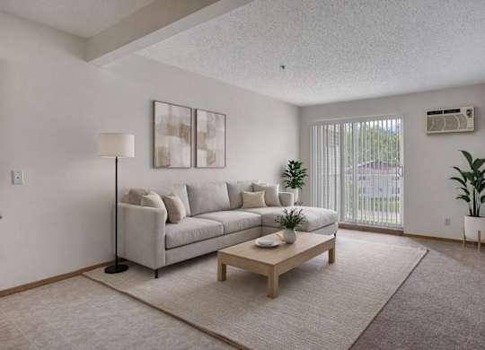 A bright living room here at Quail Ridge with soft carpet flooring, a light-gray sectional sofa, and a simple wooden coffee table on a neutral area rug. A large window with vertical blinds brings in natural light, creating a clean and relaxing vibe.