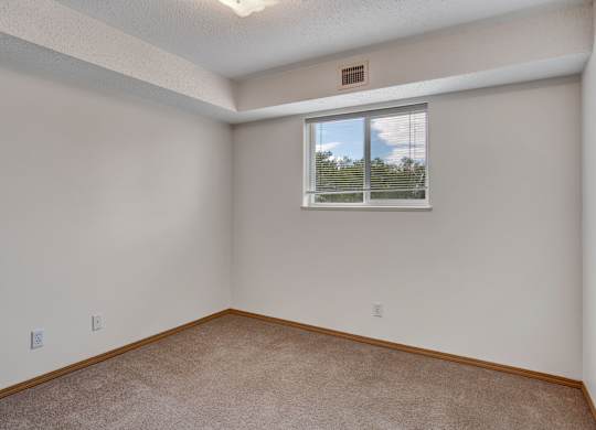 A bright, empty bedroom here at Quail Ridge with soft carpet, clean white walls, and a wide window with blinds letting in natural light. A recessed ceiling detail adds a cozy touch while keeping the space open and versatile.