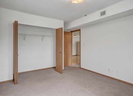 A spacious, empty bedroom here at Quail Ridge with soft carpet, clean white walls, and an open closet with built-in shelving for easy storage. A simple overhead light and warm wood trim add a bright, comfortable feel.